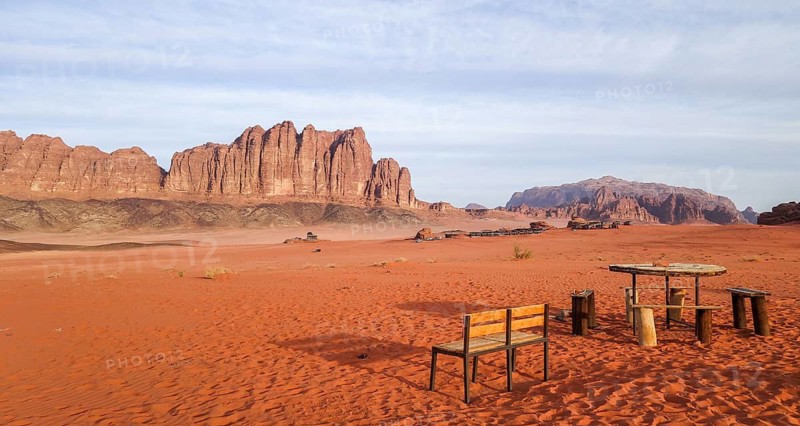 A serene view captures the essence of Wadi Rum in Jordan with its sprawling red sand and monumental rock faces. A rustic wooden picnic table and benches stand invitingly under the vast open sky