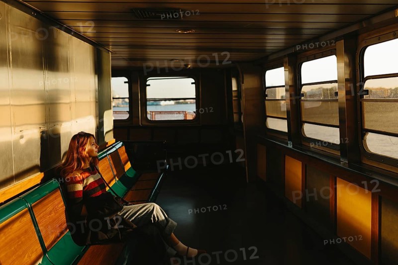 A woman sits alone on a ferry, absorbed in thought as the warm, golden light of the setting sun bathes the quiet interior, creating long shadows and a peaceful atmosphere in New York