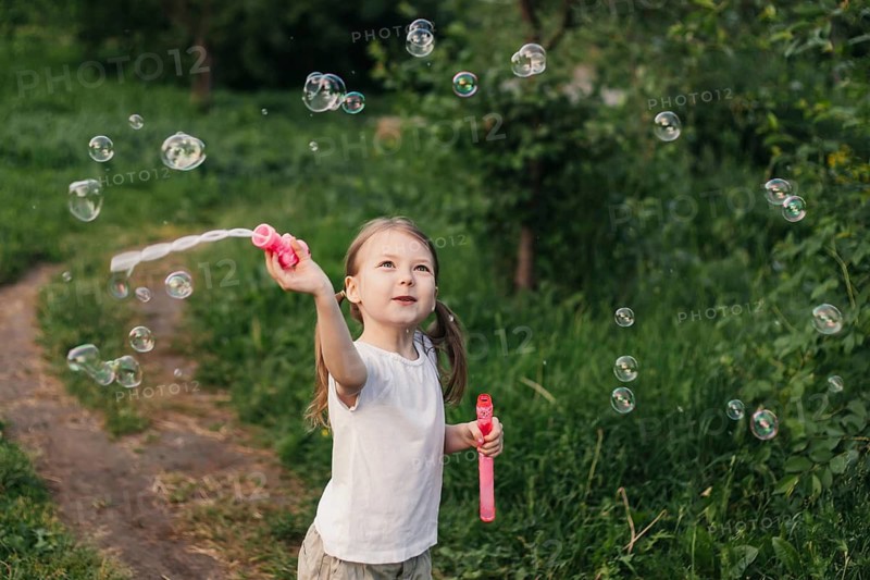 A joyful young girl delights in a summer day, playing with soap bubbles in a lush green outdoor setting. Her happiness and playfulness capture the essence of childhood joy