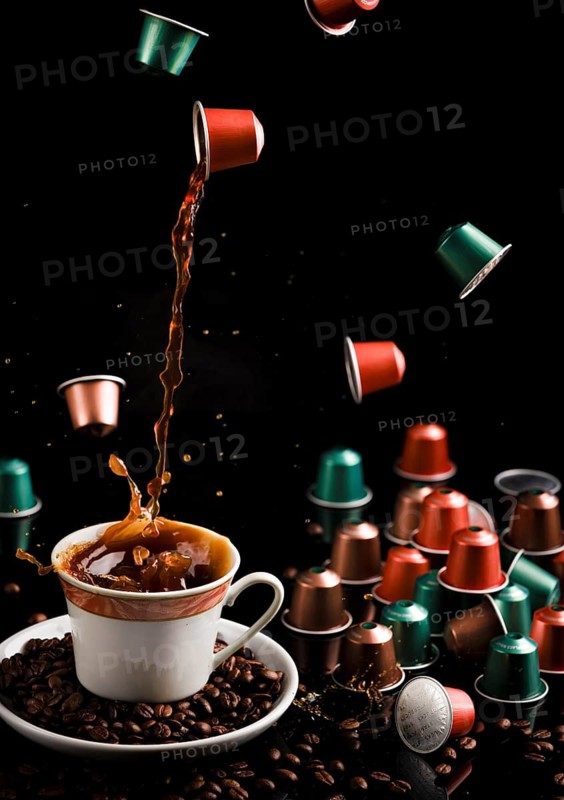Coffee flows from a floating capsule into a cup, surrounded by capsules and coffee beans