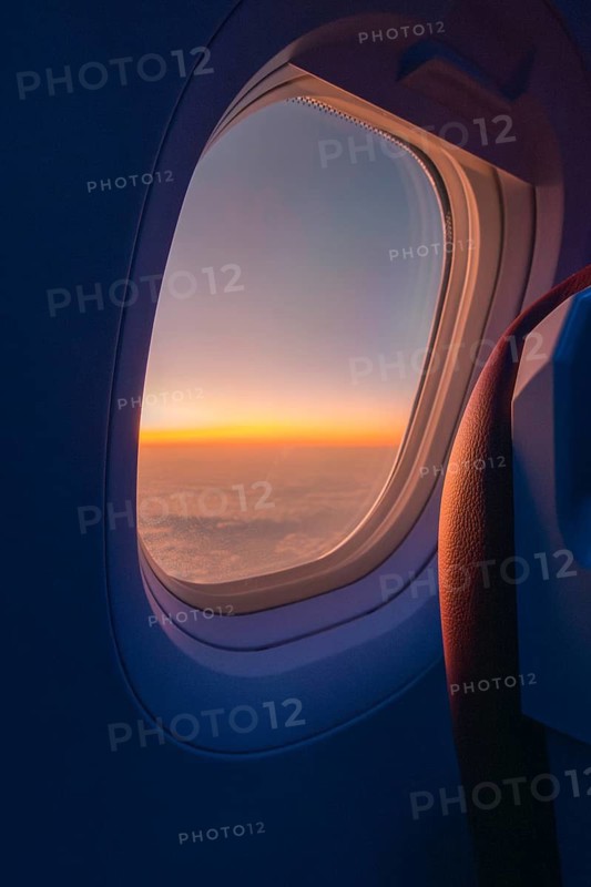 A serene sunset visible through the oval window of an airplane, showcasing a vast cloudscape tinged in hues of orange and blue