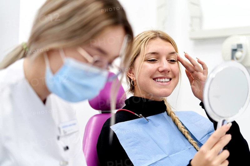 A patient is smiling and looking into a hand mirror after a successful check-up at a dental clinic. A dentist in a face mask is present, ensuring a positive experience