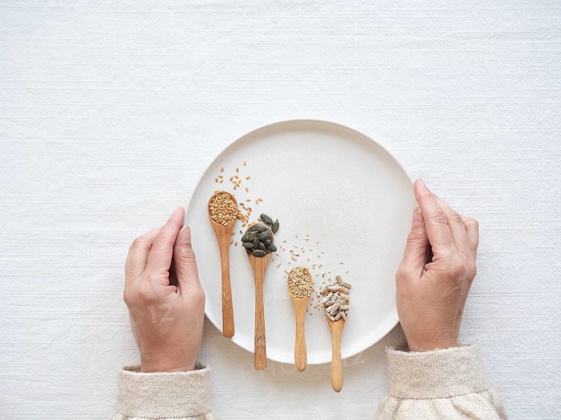 Woman holding a plate with flaxseed, pumpkin, sesame, and sunflower seeds in spoons for seed cycling