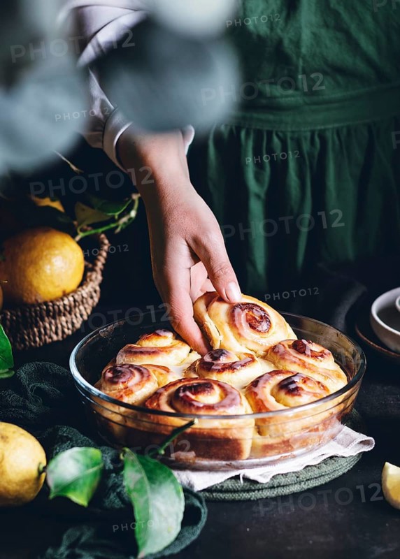 A hand reaches for freshly baked lemon rolls, perfectly golden and drizzled with glaze, in a glass dish. The setting is rustic with lemons and greenery for a fresh feel