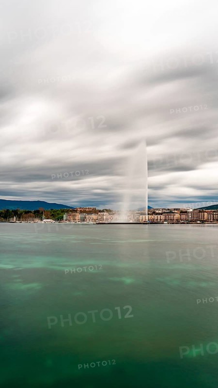 Long exposure shot of Geneva's Jet d'Eau, spectacularly soaring into the sky against a moody backdrop with the city's traditional facades lining the horizon