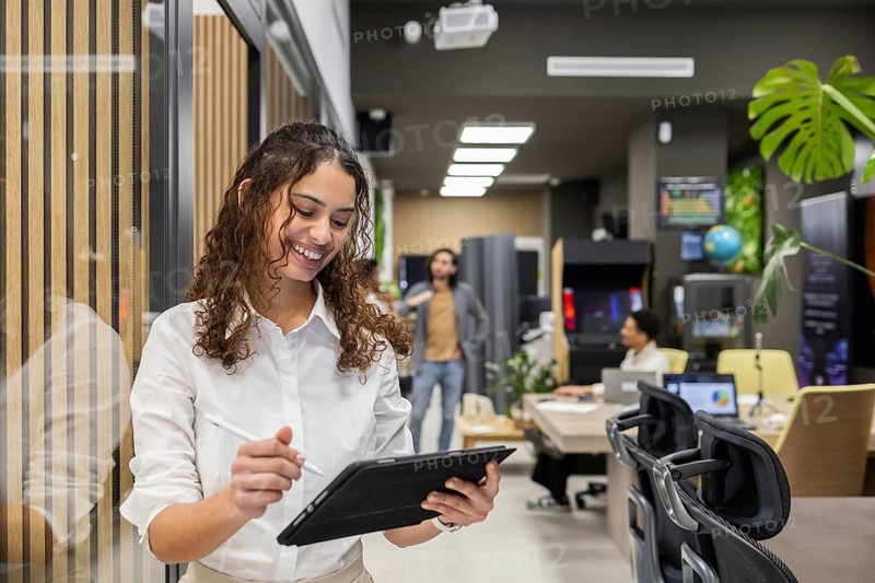 A cheerful professional in a modern office busily reviews information on a tablet. Colleagues work in the background, creating an active and collaborative work environment