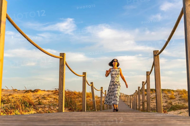 Woman wearing sunglasses and a patterned dress walking on a long wooden boardwalk, leading through the dunes to the beach, enjoying a relaxing holiday at sunset in seignosse le penon, france