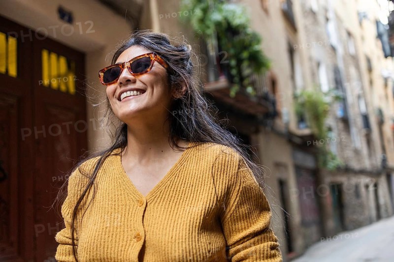 A joyful Latin woman with brown hair and wearing sunglasses grins widely while strolling through a quaint, rustic street. The sunlight enhances the beauty of the scene