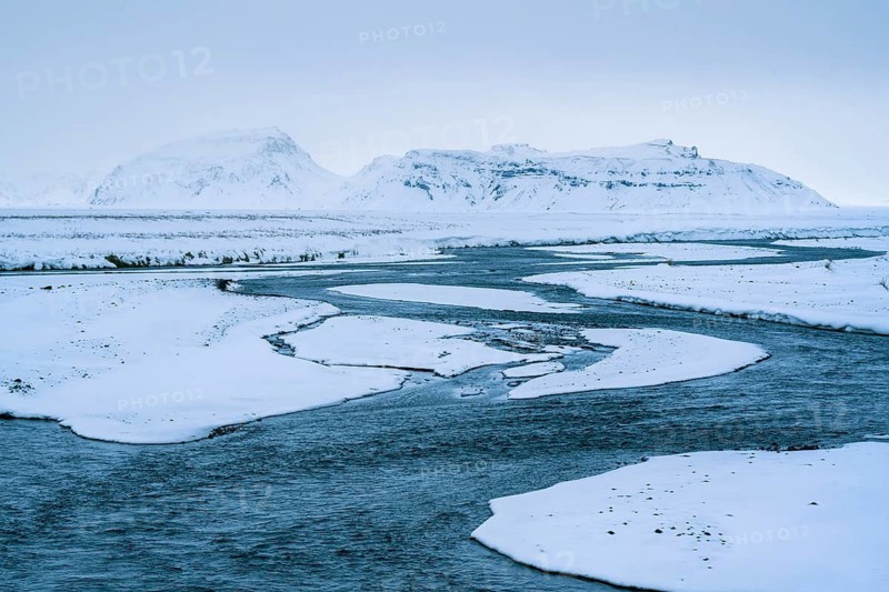 A breathtaking winter landscape of Skaftafell, Iceland, showcasing a serene river meandering through snowy plains with rugged mountains in the background, in a palette of blues and whites