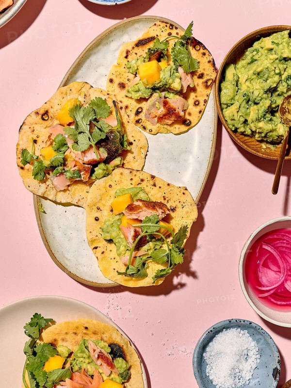 Top view of vibrant tacos prepared with sliced avocado, mango, fresh herbs, and salmon on a striking pink background alongside bowls of guacamole, pickled onions, and sea salt