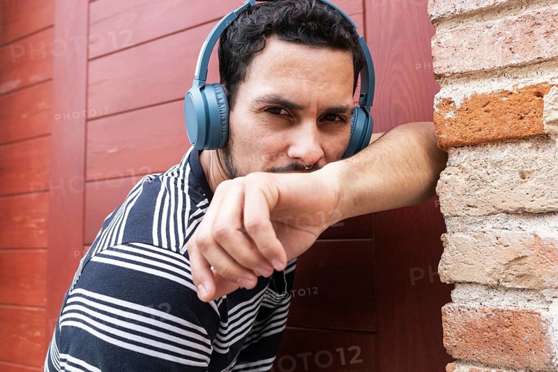 A Cuban man dancer poses confidently with headphones, wearing a striped shirt. He's leaning against a rustic red wall, expressing a sense of rhythm and style