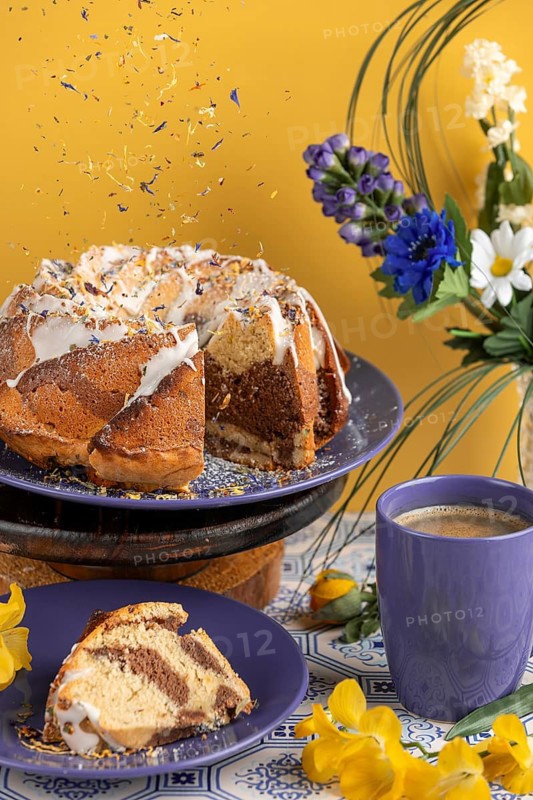 Edible flowers falling on marble cake, cake surrounded by flowers and a cup of coffee, yellow background