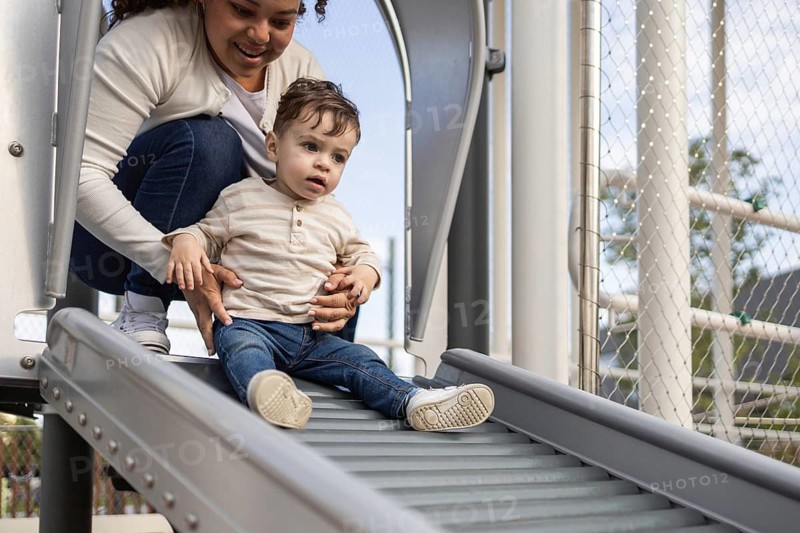 A mother and her baby enjoy a sunny day at the park. The mother helps her baby down a slide, both smiling and spending quality time outdoors in a cheerful, natural setting