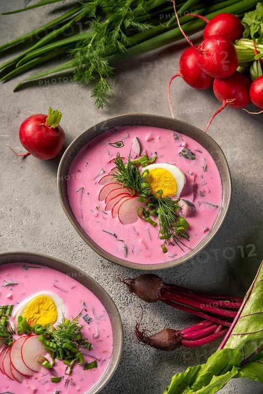 A bowl of vibrant cold beetroot soup, topped with slices of hard-boiled egg, fresh dill, and radish. Surrounding the dish are fresh greens and whole beetroots