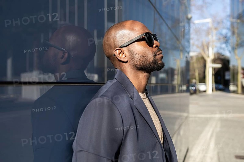 A cuban man in sunglasses and a blazer leans against a reflective building. The urban setting and confident pose create a modern and stylish atmosphere in the cityscape