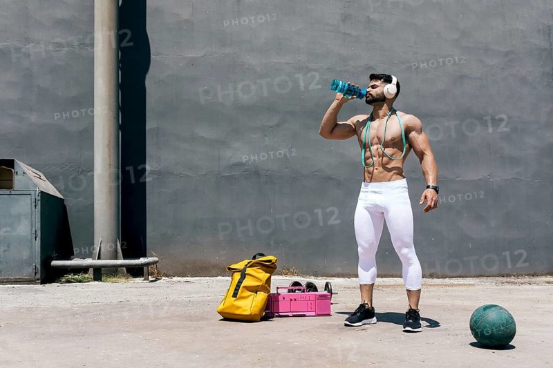 Muscular man hydrating with a water bottle after exercising outdoors, wearing white leggings, headphones, and holding resistance bands, with fitness gear scattered nearby on sunny pavement