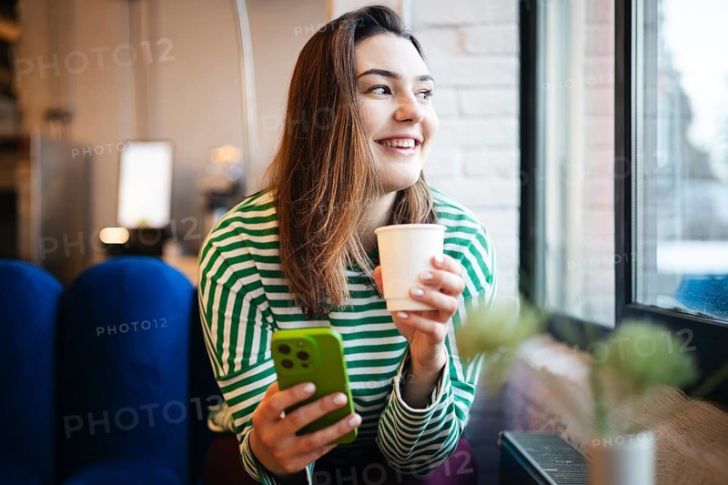 A woman in a striped shirt enjoys a coffee while using a smartphone at a cafe window, showcasing a relaxed, technology-aided break