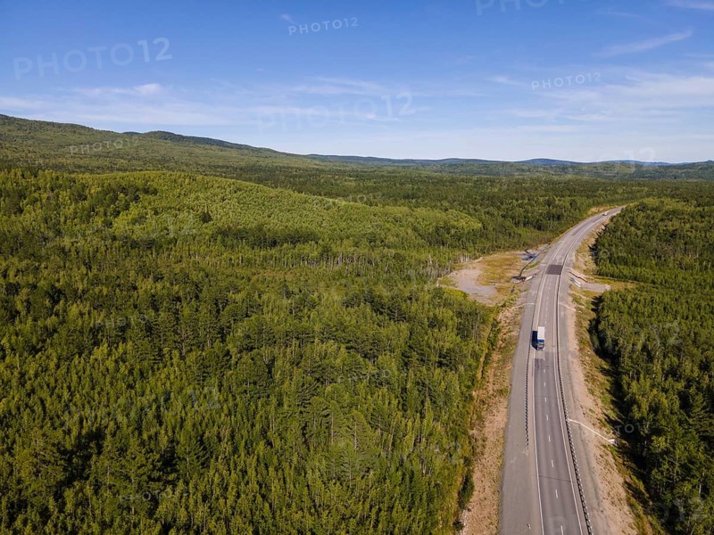 A long, winding road cuts through the dense green forests of Vladivostok, Russia. The landscape showcases the abundant nature, with trees stretching into the distance under a clear blue sky, inviting exploration