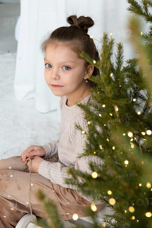 A young girl looks at the camera with soft eyes, seated beside a beautifully adorned Christmas tree. She holds a string of twinkling lights, adding to the festive atmosphere