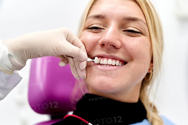 A dental professional in a clinic carefully matches tooth color for a smiling female patient, ensuring a perfect fit for restoration. The patient appears relaxed and satisfied