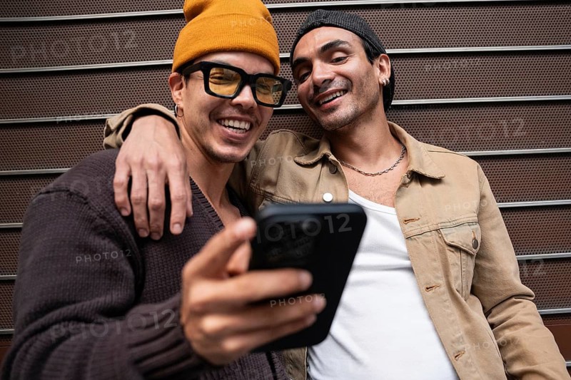 A Colombian gay couple smiles warmly as they look at a phone together. They are casually dressed, sharing a joyful moment outdoors in a relaxed setting