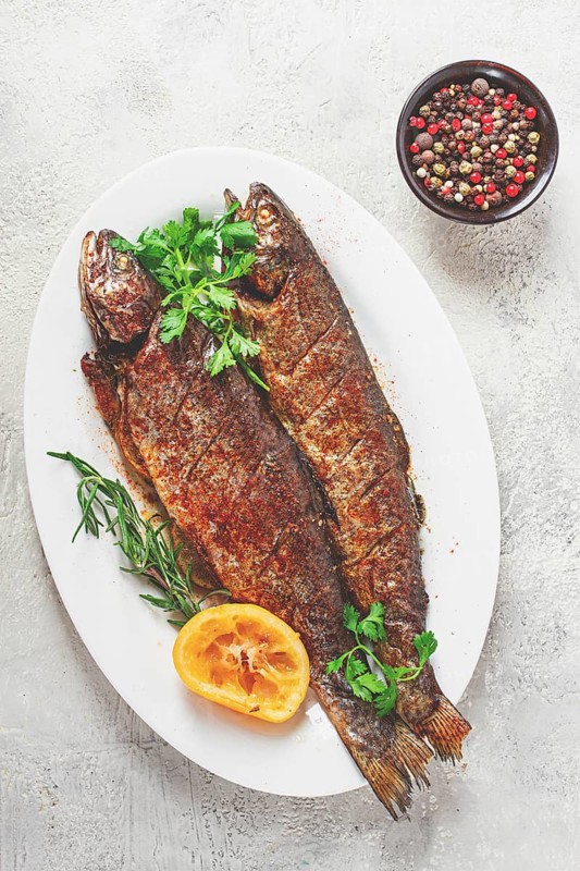 Two grilled fish are placed on a white dish. Fresh lemon slices and herbs are arranged beside them