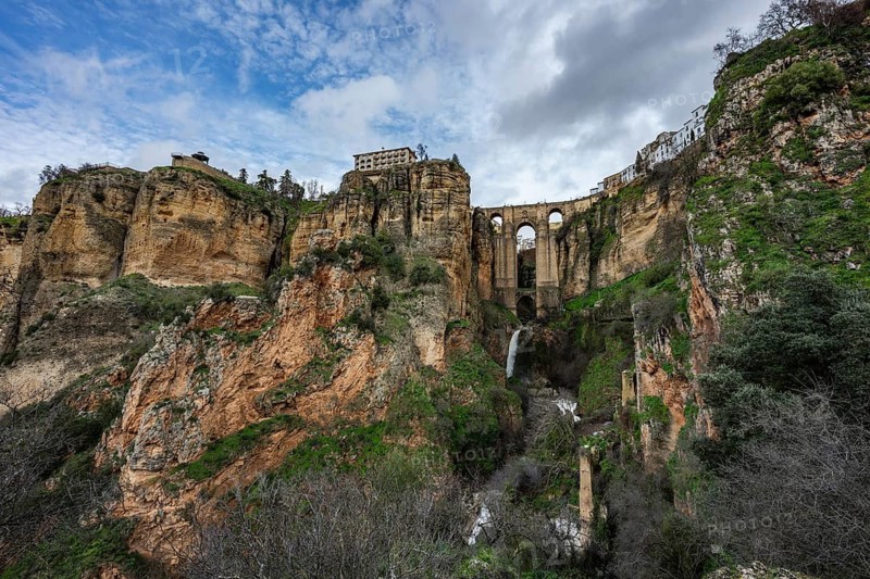 Wide angle view from the bottom of the gorge featuring the massive arches of Puente Nuevo bridge and a small waterfall in Ronda, Spain