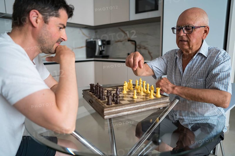 A mature father and his adult son spend quality time together at home by playing chess. This image captures a meaningful intergenerational bonding moment