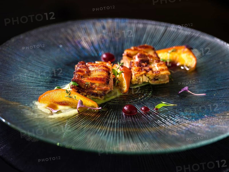 A gourmet dish featuring confit pork paired with fresh, juicy peaches and garnished with micro greens, elegantly presented on a textured glass plate