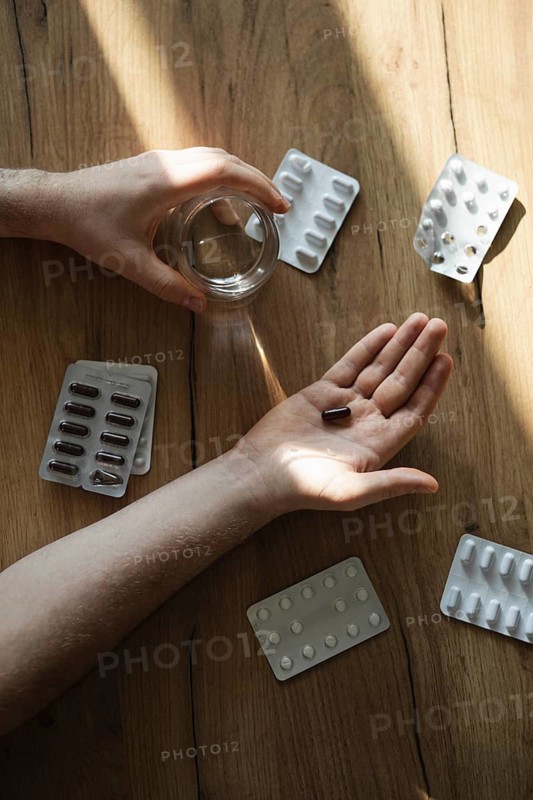 A man's hand holds a dark capsule over a wooden table, surrounded by various blister packs. A glass of water is nearby, capturing a moment of daily routine