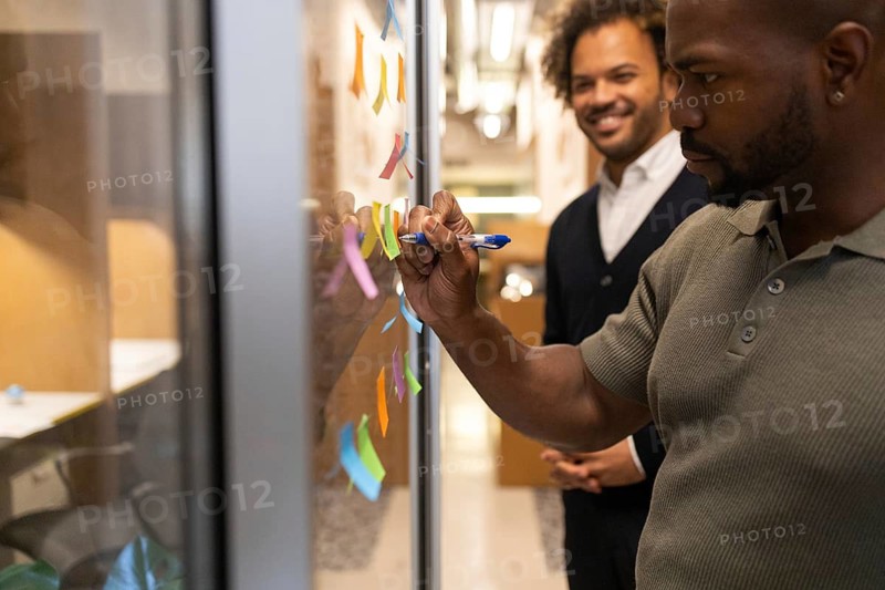 Professionals collaborate in an open coworking space in Barcelona. One person writes on glass with colorful sticky notes, while another observes, creating a dynamic team atmosphere