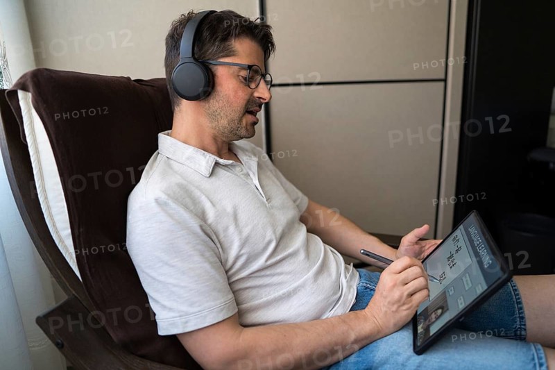 A man sits comfortably at home, focused on his tablet as he studies Mandarin online. Wearing headphones, he is engaged in a virtual language class, enhancing his skills