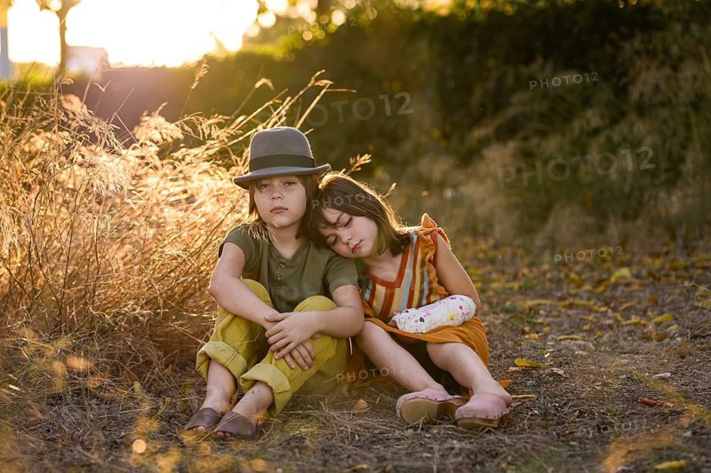 Two children sit together in a sunlit field, forming a heartwarming scene of sibling closeness. The golden hour light creates a tranquil and nostalgic atmosphere