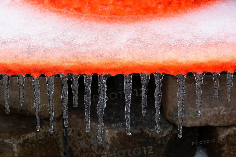 Close-up of icicles hanging from edge of flat base footing on orange fluorescent rubber traffic and construction safety cone after freezing rain in late winter, Quebec, Canada