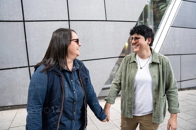 A joyous moment captured of a curvy lesbian couple, smiling and holding hands on a city street. They are casually dressed, showcasing a candid expression of love and connection