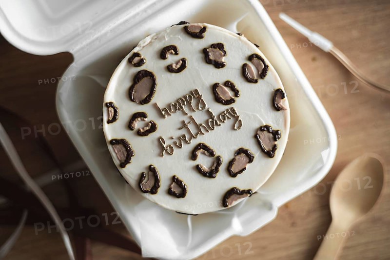 Top view of a beautifully designed Bento birthday cake inside a white takeout box, featuring elegant Happy Birthday icing surrounded by chocolate decorations