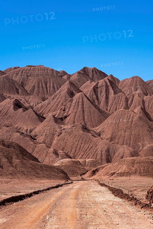 A striking image showcasing a solitary dirt road meandering through the dramatic, eroded mountain terrain in La Puna, Argentina, against a clear blue sky