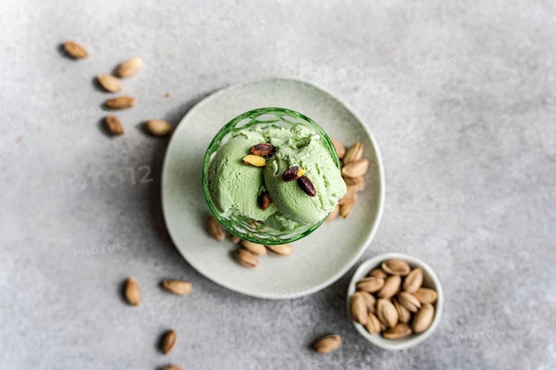 Two scoops of creamy pistachio ice cream sit in a glass bowl, garnished with whole pistachios. The dish is placed on a white plate with scattered nuts, creating a tempting dessert setting