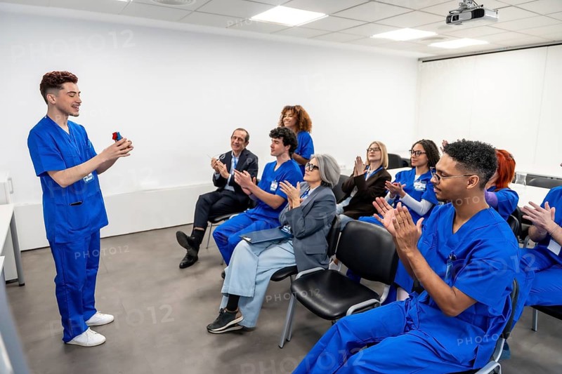 Young medical professional presenting a device during a healthcare seminar, with diverse colleagues in blue scrubs and supervisors applauding during the educational training session