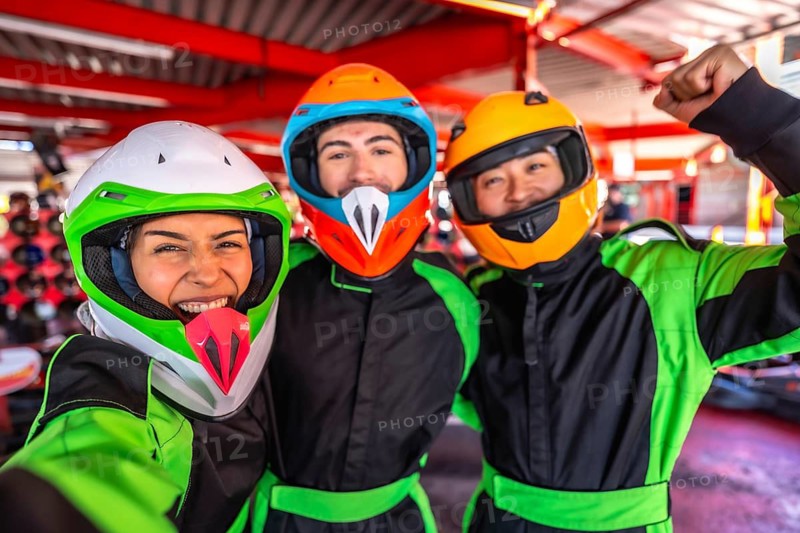Three friends wearing helmets and racing suits enjoying a day at the go kart track, capturing a fun selfie moment, celebrating their racing experience