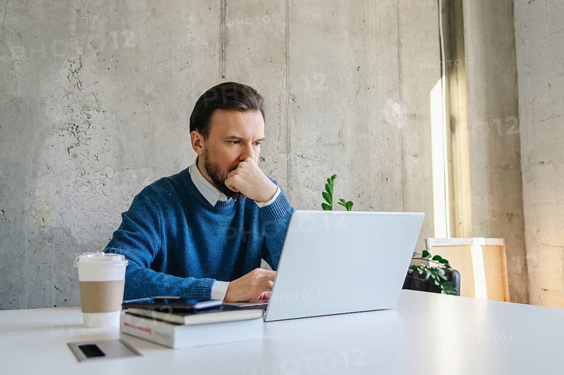 A professional man sits at a desk, intently focused on his laptop. He wears a blue sweater and has a coffee cup, books, and plant nearby, creating a modern office vibe