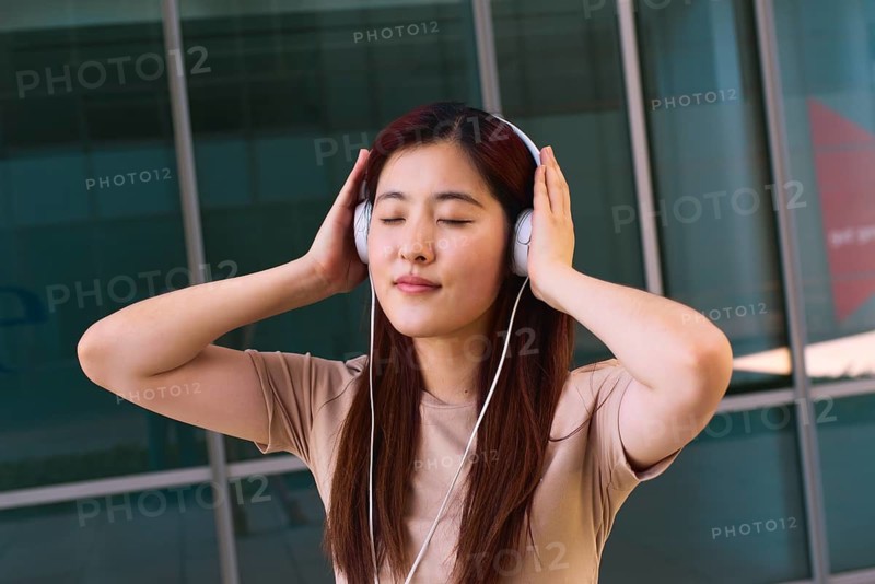 A young woman stands outside a modern building, eyes closed, fully immersed in music. She wears white headphones and a beige top, radiating calmness and relaxation