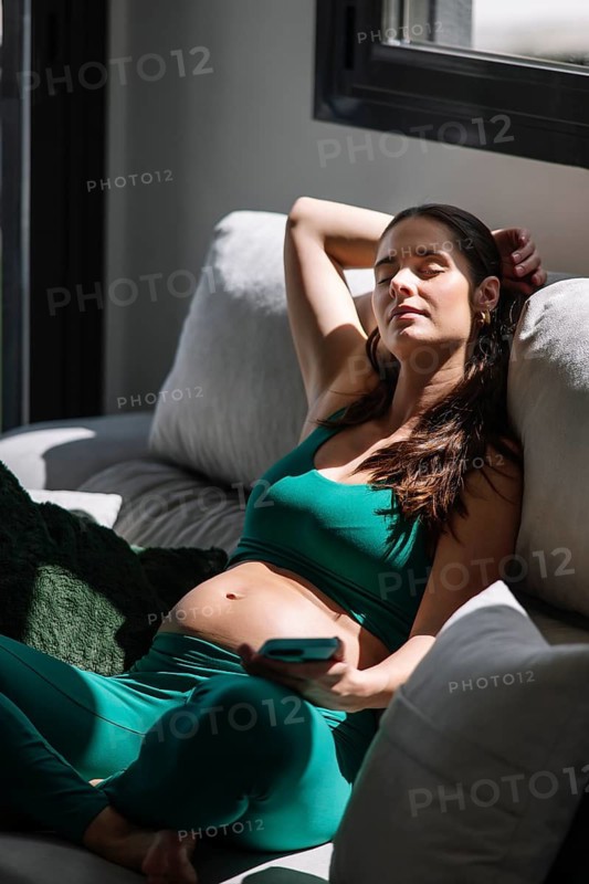 A pregnant woman relaxes peacefully on a couch, wearing a comfortable yoga outfit. Sunlight filters through the window, emphasizing her calm and serene demeanor