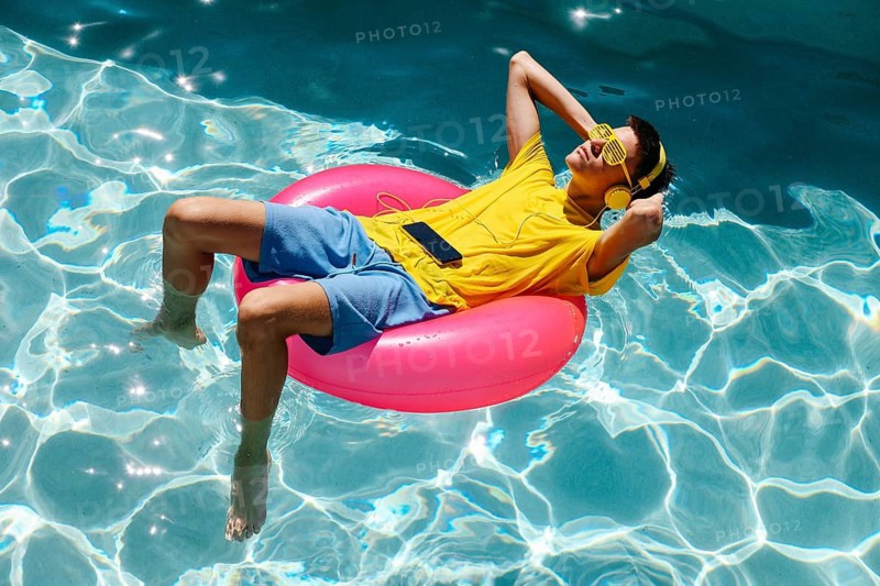 A young teenage boy relaxes on a pink pool inflatable, wearing a yellow shirt and sunglasses. He's enjoying music through headphones under the bright sun