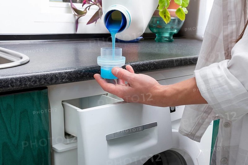 A Gen-Z girl pours vibrant blue liquid detergent into a cap in preparation for a laundry cycle, illustrating a clean and fresh start to household chores in a cozy home setting