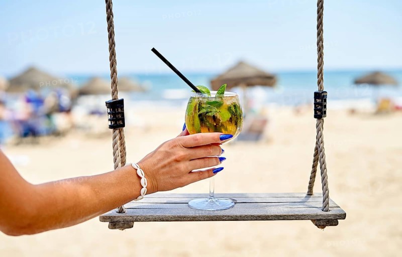 A colorful drink with mint and a straw is held from a swing overlooking a sandy beach. Sunbathers relax under straw umbrellas while waves lapping in the distance create a serene atmosphere
