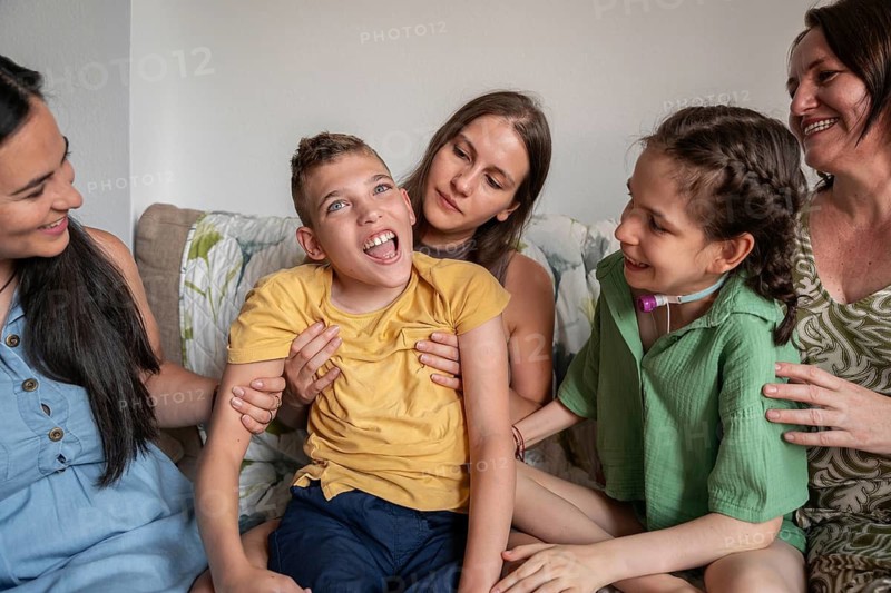 A joyful family shares a tender moment with a girl needing a tracheostomy and a boy with cerebral palsy, highlighting love and support in a diverse and caring environment