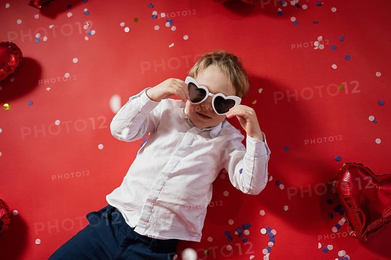 A young boy in a white shirt wears heart-shaped sunglasses, celebrating Valentine's Day on a red background with confetti and balloons