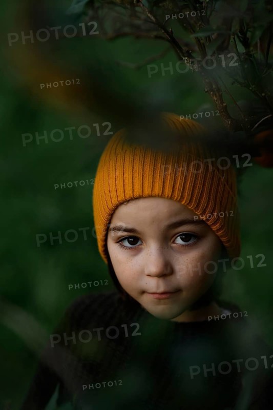 A child, wearing a cozy orange hat, peeks through lush, green foliage, creating a sense of curiosity and exploration in a tranquil forest setting