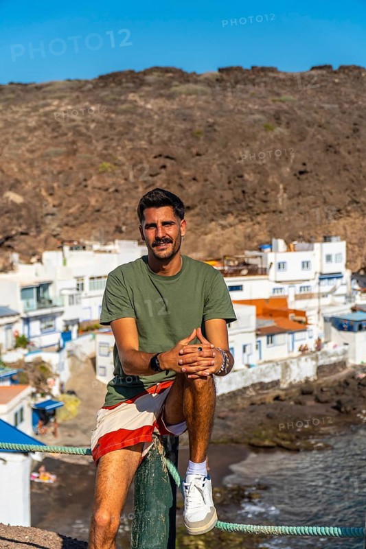 Young man sitting on a wooden pole enjoying the view of the colorful houses of tufia, a coastal village in gran canaria, canary islands, spain